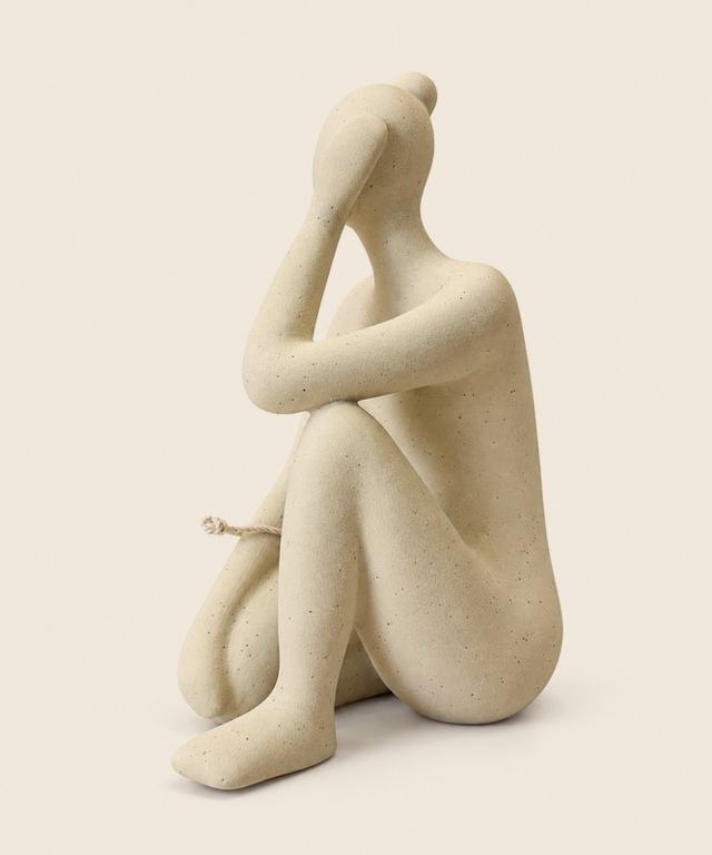 Escultura color arena de una figura sentada que abraza sus rodillas.