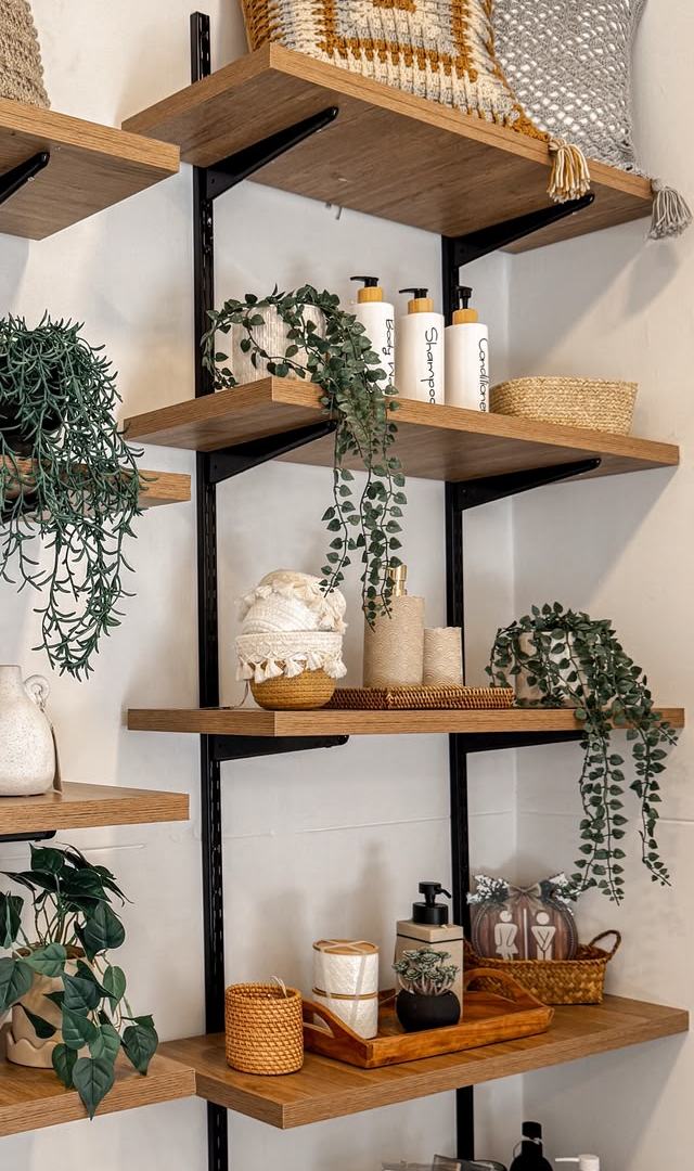 Repisas de madera con cojines tejidos, una cesta con borlas, dispensadores de cerámica y plantas colgantes.
