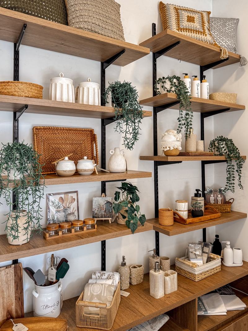 Pared de estanterías de madera en Casa Luma con tarros de cerámica blanca, una bandeja de rattán, plantas colgantes y cojines tejidos.