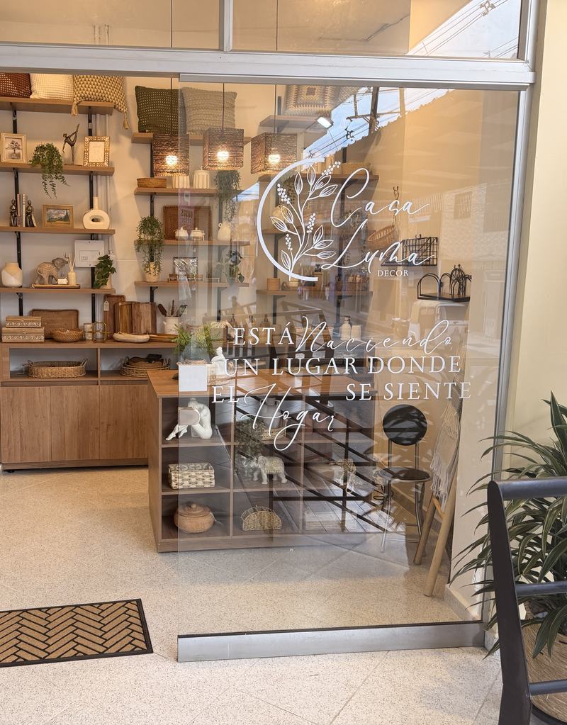 Vitrina de Casa Luma en Guarne, con el logo y la frase «Está naciendo un lugar donde el hogar se siente» sobre el vidrio, y estanterías de madera al fondo.