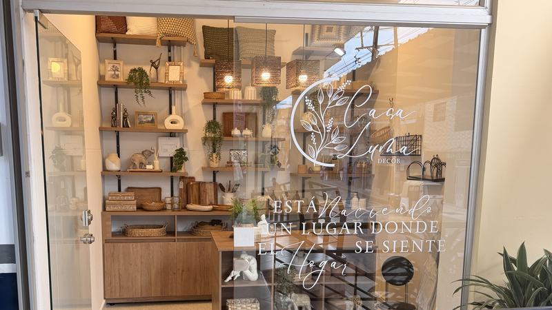 Fachada de Casa Luma en Guarne: puerta de vidrio abierta, el logo en la vitrina y estanterías de madera con cestas, cojines y cerámica.
